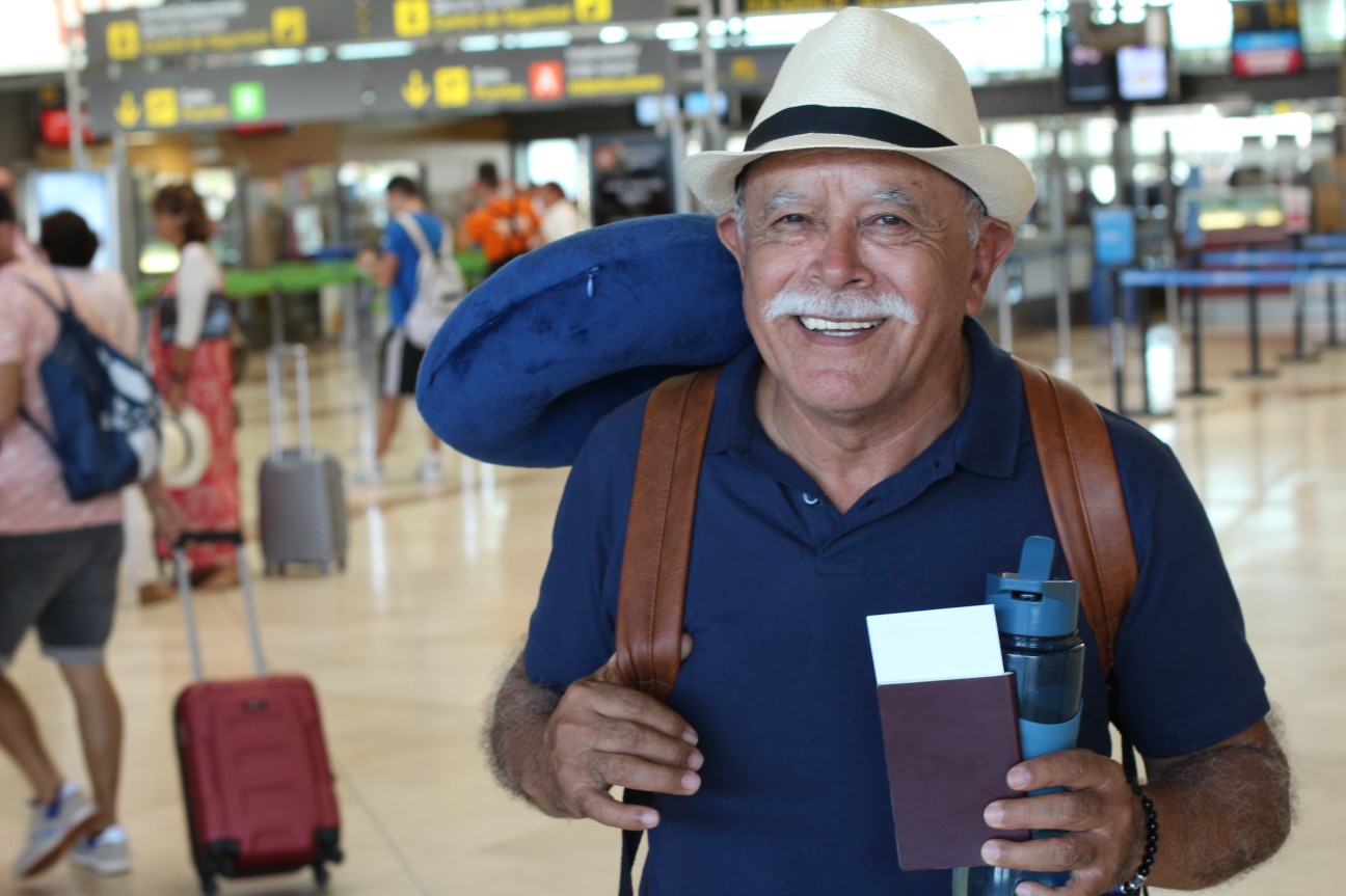 Idodo de chapeu e mochila, sorrindo com um bilhete de avião na mão, em um aeroporto