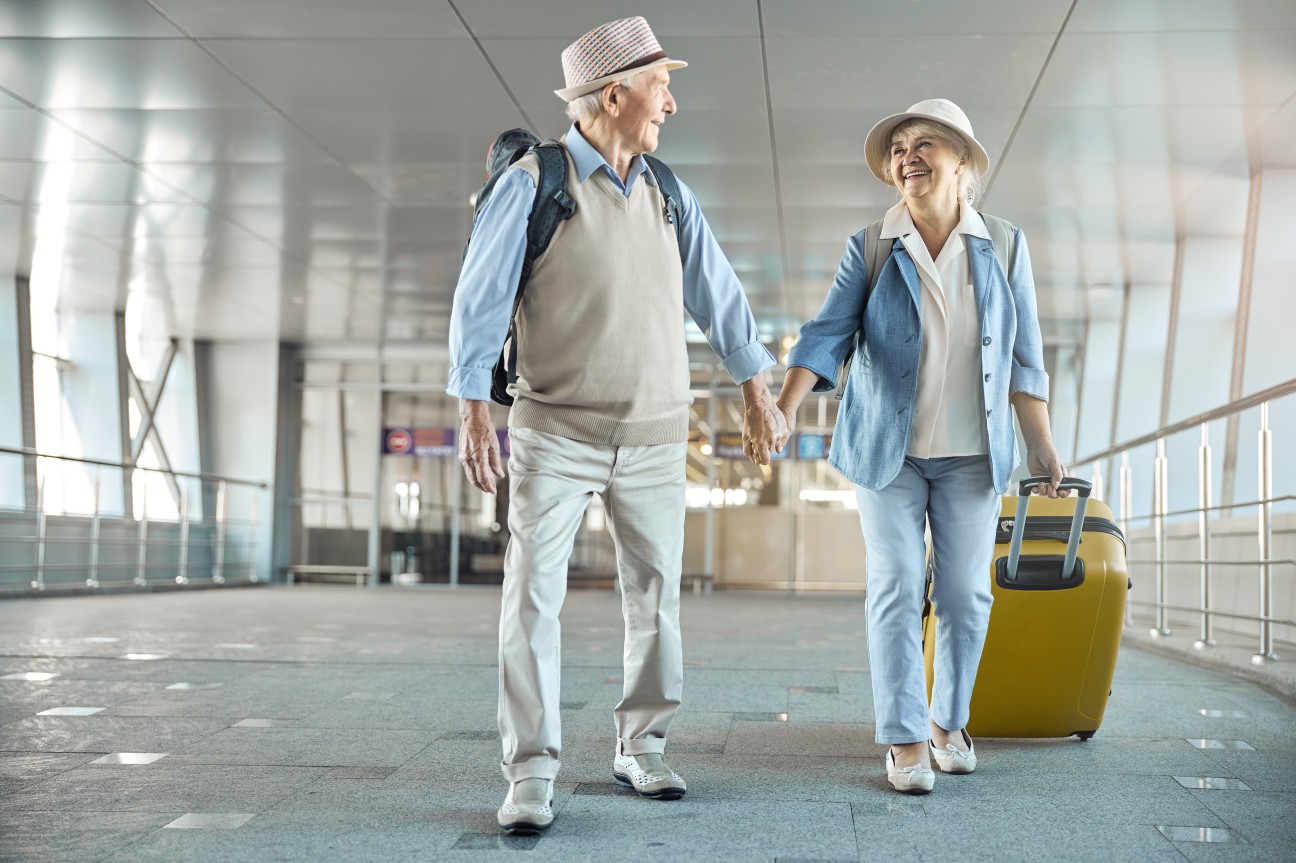 Casal de idosos com chapéu, mochila nas costas e mala, sorrindo em um aeroporto